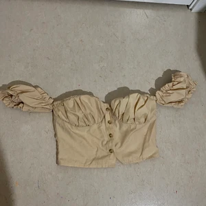 Blus/topp med knäppning strl 34 - En liten söt beige blus med knäppning där fram och off shoulder från prettylittlething. Aldrig använd. Tajt och croppad i passform!