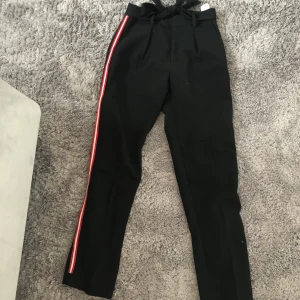Kostymbyxor svarta och röda Storlek 32 - Snygga kostymbyxor med knytning upp till och röda och vita linjer på sidan. Använda endast ett fåtal gånger.