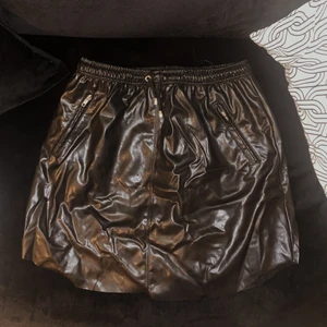 Svart skinnkjol med dragsko i midjan - Svart skinnkjol (ej äkta skinn) med dragsko i midjan från NA-KD. Två fickor med dragkedja. Stretchigt material. Avklippt modell. Storlek 42. Gott skick.