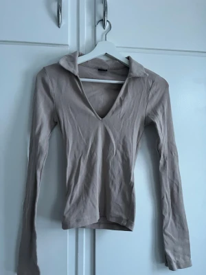 Beige långärmad polotopp - Trendig beige långärmad topp med krage och djup v-ringning. Tillverkad i mjukt bomullsmaterial och har en slim passform som framhäver siluetten. Perfekt att styla med jeans eller kjol för en clean look.