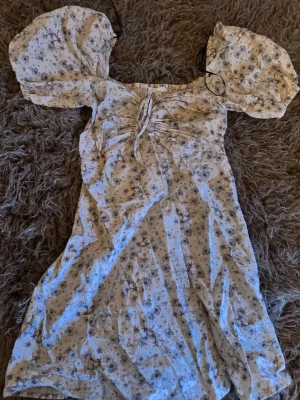 Blommig kortklänning med puffärm - Söt kortklänning i ljus färg med småblommigt mönster och puffiga kortärmar. Klänningen har snörning vid bysten och en lätt v-ringad halsringning. Perfekt för sommarens alla tillfällen.