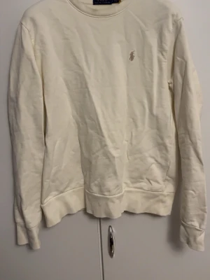 Beige sweatshirt från Polo Ralph lauren - Klassisk vit sweatshirt från Ralph Lauren med rund halsringning och diskret broderad logga på bröstet. Tröjan har ribbade muddar vid ärmslut och nederkant. Perfekt för en clean och stilren look.