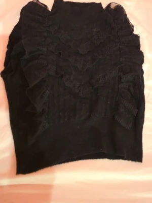 Svart topp med volanger och spets - Snygg svart topp med höghalsad design, dekorerad med volanger och spetsdetaljer framtill och längs ärmarna. Ribbad struktur ger en extra touch och toppen har en croppad passform. Perfekt för dig som gillar unika detaljer och en trendig look.