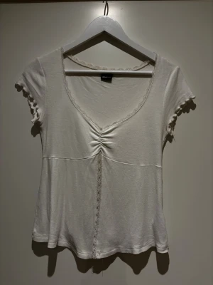 Vit topp med spets och rynk från Gina Tricot - Söt vit topp från Gina Tricot med korta ärmar, v-ringning och spetsdetaljer längs halsringning och mittsöm. Toppen har rynk framtill och en dekorativ volangkant vid ärmslut.