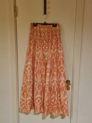Långkjol med orange mönster - Säljer en flowy långkjol med vitt och orange mönster. Kjolen har bred resår i midjan och volangdetaljer som ger en bohemisk känsla. Perfekt för dig som gillar färg och mönster. Materialet känns lätt och luftigt.