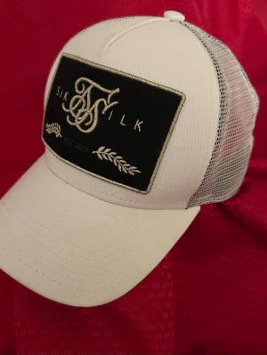 Vit truckerkeps från SikSilk - Snygg vit truckerkeps från SikSilk med svart patch framtill och broderad logga. Kepsen har böjd skärm och mesh på sidorna och bak för extra ventilation. Perfekt accessoar för en streetwear-look.