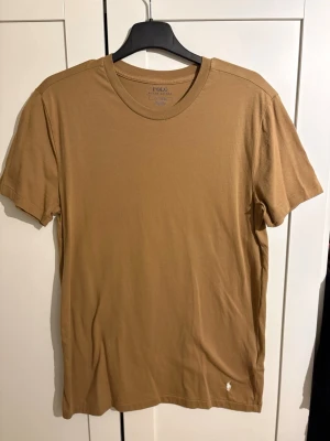 Beige t-shirt från Polo Ralph Lauren - Snygg beige t-shirt från Polo Ralph Lauren med klassisk rund halsringning och liten broderad logga vid nederkanten. Tillverkad i mjuk bomull som känns skön mot huden. Perfekt för en avslappnad och stilren look.