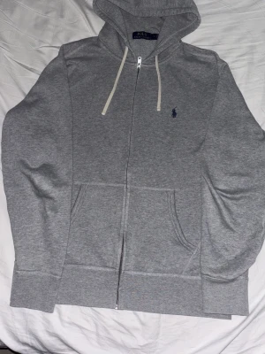 Grå hoodie från Polo Ralph Lauren - Klassisk grå hoodie med dragkedja från Polo Ralph Lauren. Mycket bra skick och är i storlek M. Den är äkta vilken man kan se genom att skanna QR koden. Mycket snygg o skön, passar till allt!