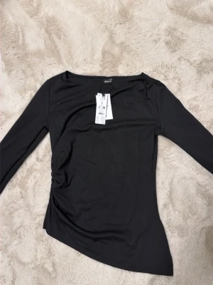 Svart långärmad topp  - Svart långärmad topp från NA-KD med asymmetrisk nederkant och lätt rynkdetalj på sidan. Tunn och stretchig kvalitet, perfekt att styla med jeans eller kjol. Enkel men trendig design med rund halsringning.