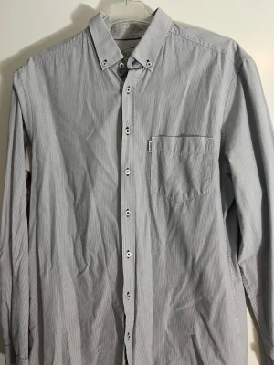 Randig skjorta från Dobber - Klassisk långärmad skjorta från Dobber med tunna vertikala ränder i vitt och grått. Skjortan har knappar hela vägen, bröstficka och en liten etikett med Dobber-logga. Perfekt till jeans eller chinos för en clean look.