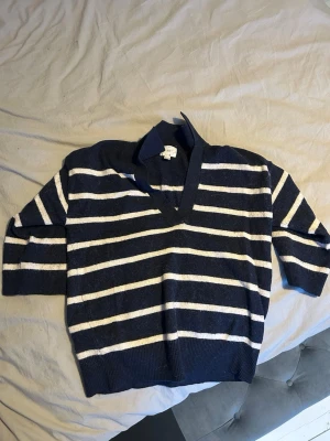 Marinblå randig stickad tröja H&M - Snygg marinblå stickad tröja från H&M med vita horisontella ränder. Tröjan har V-ringning och krage, samt ribbade muddar vid ärmslut och nederkant. Perfekt för en avslappnad men stilren look.