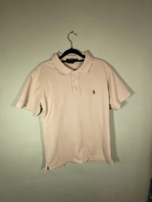 Beige pikétröja från Polo Ralph Lauren - Klassisk beige pikétröja från Polo Ralph Lauren i custom slim fit. Tröjan har korta ärmar, krage och knappar framtill samt den ikoniska broderade loggan på bröstet.  Perfekt till jeans eller shorts för en clean och stilren look. Storlek S/M.