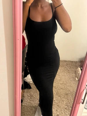 Svart långklänning med smala axelband - En stilren svart långklänning med smala axelband och figurnära passform. Klänningen har en enkel och klassisk design som passar perfekt till många olika tillfällen. Materialet känns mjukt och följsamt mot kroppen.