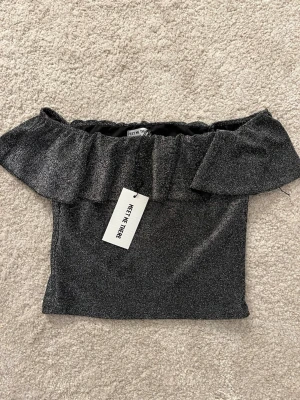 Glittrig offshoulder topp Meet Me There - Supersnygg glittrig svart offshoulder topp från Meet Me There. Toppen har volang upptill och är perfekt för dig som vill sticka ut. Materialet är stretchigt och sitter snyggt på kroppen. Perfekt att styla med jeans eller kjol.