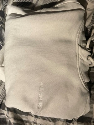 Vit hoodie från J.Lindeberg - Stilren vit hoodie från J.Lindeberg med broderad logga på bröstet. Klassisk känguruficka och dragsko i huvan. Perfekt för en avslappnad och trendig look. Köptes ny för 1700kr. Kan passa om man har storlek M också.