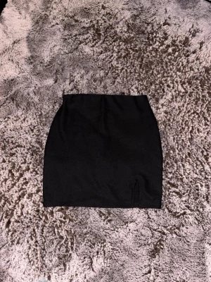 Svart kortkjol med slits - En stilren svart kortkjol med liten slits framtill. Kjolen har en enkel och tight passform som passar perfekt till många olika outfits. Perfekt basplagg för garderoben.
