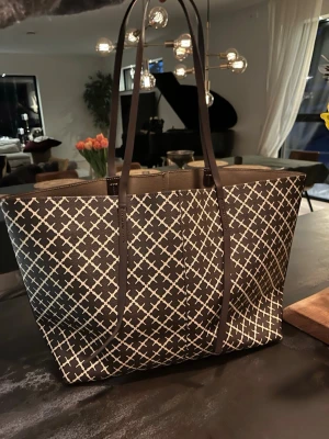 Brunvit shoppingväska By Malene Birger - Snygg shoppingväska från By Malene Birger i brunt och vitt med ett grafiskt rutigt mönster. Väskan har långa handtag och är tillverkad i tålig syntet. Perfekt för dig som vill ha plats för allt och samtidigt se trendig ut.