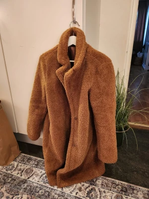 Brun oversized teddyjacka - Mysig och trendig brun teddyjacka med oversized passform och hög krage. Jackan har långa ärmar och stängs med knappar framtill. Perfekt för att hålla värmen under kyliga dagar och ger en cool look till din outfit.