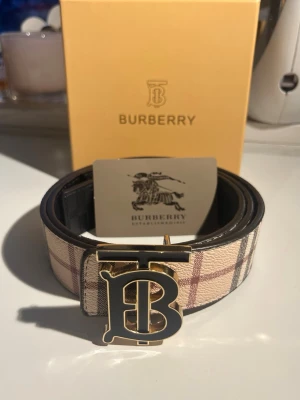 Burberry bälte med monogramspänne - Snyggt Burberry bälte i klassiskt beige rutmönster med stort guldfärgat och svart TB-monogram som spänne. Tillverkat i läder med exklusiv känsla och ikonisk design. Perfekt accessoar för att lyfta din outfit med en lyxig touch.