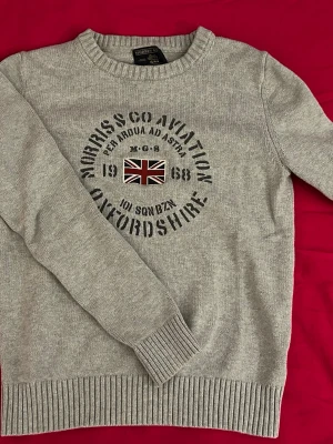 Grå stickad tröja Morris & Co Aviation - Grå stickad tröja med rund hals från Morris & Co Aviation. Framsidan har tryck och broderad Union Jack-flagga samt texten 'OXFORDSHIRE' och '1968'. Tröjan har ribbstickade muddar och nederkant. Perfekt för dig som gillar brittisk stil, har defekt på lappen då den gått lite ur stygn.