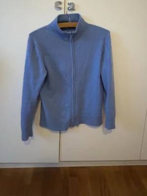 Cashmere zip - Ljusblå stickad tröja med dragkedja framtill och hög krage. Tröjan har långa ärmar och ribbade muddar vid krage, ärmslut och nederkant. Perfekt för lager-på-lager. Finns en liten defekt (bild 4)