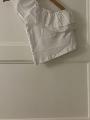 Vit oneshoulder topp med volang - Snygg vit oneshoulder topp med bred volangkant i broderad spets. Perfekt för dig som gillar en feminin och trendig stil. Toppen är ärmlös och har en luftig passform. Bara att fråga ifall du undrar något eller om du vill ha fler bilder! Pris kan diskuteras💞