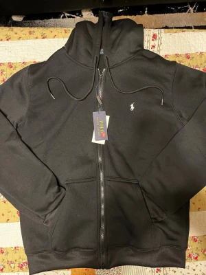 Polo Ralph Lauren hoodie  - Snygg svart hoodie från Polo Ralph Lauren med dragkedja framtill, två fickor och justerbar huva med snören. Klassisk logga broderad på bröstet. Tillverkad i mjukt material som känns skönt mot huden. Perfekt för en avslappnad och stilren look. Helt ny 