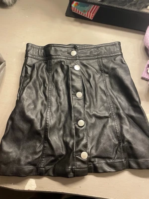 Svart kort kjol i skinnimitation - Snygg svart kort kjol i skinnimitation med silverfärgade knappar framtill. Kjolen har en rak passform och markerad midja, perfekt att styla med en crop top eller stickad tröja. Ger en cool och edgy vibe till din outfit.