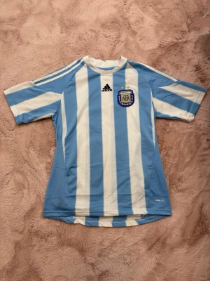 Argentina Adidas fotbollströja - Säljer en klassisk Argentina fotbollströja från Adidas med ljusblå och vita vertikala ränder, broderad AFA-logga och svarta Adidas-detaljer. Tröjan har korta ärmar och rund hals, perfekt för fotboll eller träning.
