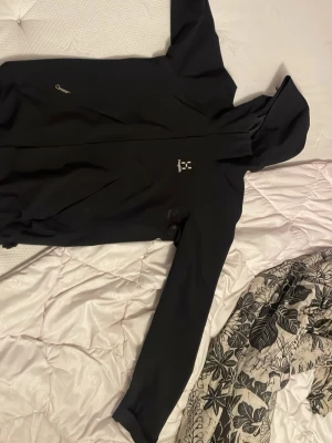 Svart vindjacka från Haglöfs med huva - Säljer en svart vindjacka från Haglöfs med hel dragkedja framtill och justerbar huva. Jackan har långa ärmar, minimalistisk design och dragsko i nederkant för extra passform. Perfekt för blåsiga dagar och outdoor-äventyr.