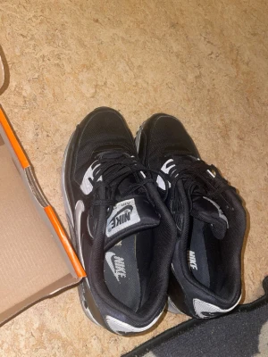 Svarta Nike Air Max sneakers - Snygga svarta Nike Air Max sneakers med vita detaljer och klassisk Nike-logga på sidan. Skorna har snörning och en bekväm platt sula med synlig Air Max-dämpning. Tillverkade i en mix av mesh och syntetmaterial för både stil och komfort.