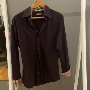 Mörklila Burberry skjorta med rutig detalj - Snygg mörklila skjorta från Burberry med klassiska rutiga detaljer på insidan av kragen och ärmsluten. Skjortan har lång ärm, knappar framtill och en broderad Burberry-logga på bröstet. Perfekt för dig som gillar stilrena plagg med lyxig touch. Har ett lite hål vid armhålan 