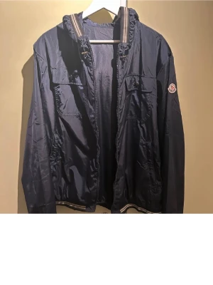 Moncler vindjacka - Snygg marinblå vindjacka från Moncler med huva och dragkedja. Jackan har två bröstfickor med lock, ribbade detaljer vid krage och midja samt Moncler-logga på ärmen. Perfekt för blåsiga dagar.
