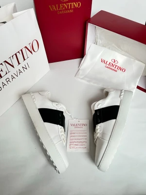 Valentino Garavani vita sneakers - Snygga vita sneakers från Valentino Garavani med en bred svart rem över sidan. Skorna har snörning, platt sula och rund tå. Tillverkade i skinn för en lyxig känsla och stilren look. Perfekta för dig som vill ha något trendigt och exklusivt.