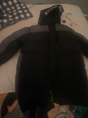 Svart dunjacka från Zavetti Canada - Säljer en svart dunjacka från Zavetti Canada med huva och logga på insidan samt huvan. Jackan har en sportig look med breda paneler och dragkedja framtill. Perfekt för kalla dagar när du vill hålla dig varm och se snygg ut.