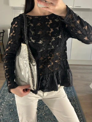 Svart spetsblus med volangkant - Trendig svart blus i spets med långärmad design och volangkant nedtill. Blusen har ett transparent blommönster och rund halsringning. Perfekt att styla med både jeans och kjol för en cool look.