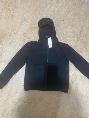 C.P. Company Goggle Hoodie – Svart – S – Ny med tags - Säljer en svart hoodie från C.P. Company med dragkedja och ikoniska inbyggda glasögon i huvan. Hoodien har två stora fickor framtill och ribbade muddar. Perfekt för dig som vill sticka ut med en unik streetwear-look.
