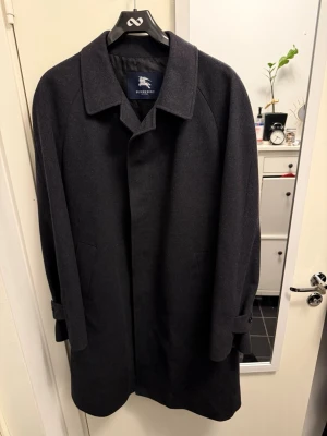 Mörkgrå ullkappa från Burberry - Stilren mörkgrå kappa från Burberry i ull med klassisk krage och knäppning framtill. Kappan är i knä längd, samt justerbara band vid ärmsluten. Perfekt för dig som vill ha en tidlös och elegant look. Använd under 10 gånger skick 9,5 köpt i madrid. Säljer pga stor på mig som vanliga fall har 46 i storlek skulle säga att den passar storlek 50+ beroende hur ni vill den ska sitta🤟