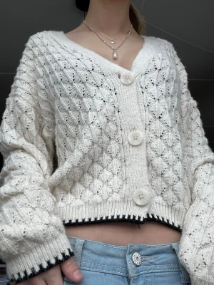 Vit stickad kofta med stora knappar - Säljer en krämvit stickad kofta med stora vita knappar och ett snyggt hålmönster. Koftan har v-ringning, ribbade muddar och svart detalj vid ärmslut och nederkant. Perfekt för lager på lager och ger en chill vibe till din outfit.