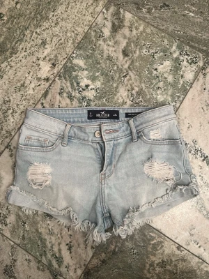 Ljusblå slitna jeansshorts Hollister - Säljer ett par ljusblå low-rise jeansshorts från Hollister med slitna detaljer och fransig kant. Shortsen har klassisk femficksdesign och knappgylf. Perfekta för sommaren.