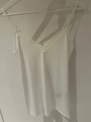 Vitt spetslinne från Pieces i storlek S - Säljer ett vitt linne från Pieces med tunna axelband och vacker spetsdetalj längs urringningen. Materialet är mjukt och följsamt, perfekt för att bära under kläder eller som ett snyggt lager. Enkel och stilren design som passar till mycket.