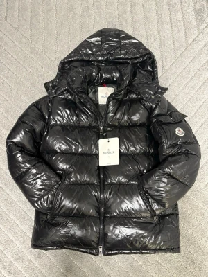 Svart dunjacka från Moncler med huva - Snygg svart puffer dunjacka från Moncler med glansig finish och stor huva. Jackan har dragkedja framtill, quiltad design och en Moncler-logga på ärmen. Perfekt för kalla dagar och riktigt trendig look.