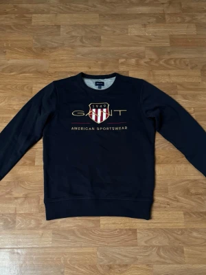 Mörkblå tracksuit från GANT - Säljer ett par mörkblå mjukisbyxor från GANT med broderad logga och text på benet. Byxorna har snörning i midjan och är gjorda i mjukt bomullsmaterial. Perfekta för chill eller sportiga dagar.