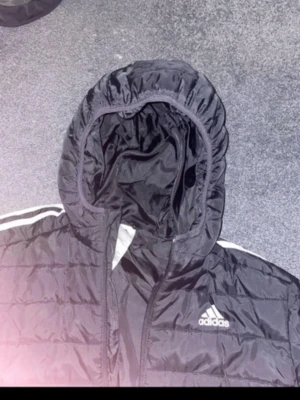 Svart dunjacka från Adidas med huva - Säljer en svart dunjacka från Adidas med klassiska vita ränder på axlarna och logga på bröstet. Jackan har huva, dragkedja framtill och quiltad design som ger en sportig vibe. Perfekt för kalla dagar när du vill hålla stilen.