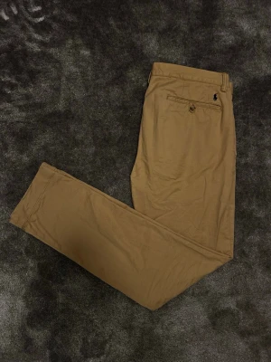 Beiga chinos från Polo Ralph Lauren - Snygga beiga chinos från Polo Ralph Lauren med klassisk rak passform. Byxorna har bälteshällor, knappstängning och den ikoniska broderade loggan ovanför bakfickan. Perfekta för en stilren och avslappnad look.