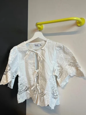 Vit blus med broderi från NA-KD - Superfin vit blus från NA-KD med broderade detaljer och knytning framtill. Blusen har vida trekvartsärmar och vackra spetsinslag som ger en bohemisk känsla. Perfekt till jeans eller kjol för en trendig look.