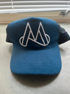 Blå truckerkeps från Maggiore - Snygg blå truckerkeps från Maggiore med vit broderad logga framtill. Kepsen har svart mesh baktill och justerbar snapback för perfekt passform. Perfekt accessoar för en cool och avslappnad stil.