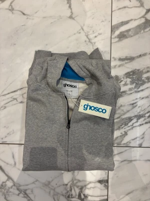 Grå zip hoodie  - Snygg grå hoodie med dragkedja framtill och huva med blått innerfoder. Tröjan har svarta detaljer på sidorna, ribbade muddar vid ärmslut och nederkant samt en liten logga på bröstet. Perfekt för en chill och avslappnad stil. Den säljs i storleken S/M/L så skriv vilken storlek du vill ha så löser jag.