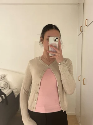 Beige kofta med rund hals - Söt beige kofta med rund halsringning och knappar framtill. Långärmad och i mjukt, tunt stickat material som passar perfekt till lager-på-lager. Enkel och stilren design som funkar till många outfits.