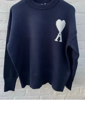 Mörkblå stickad tröja från Ami - Mörkblå stickad tröja från Ami Alexandre Mattiussi med vit hjärtlogga och bokstaven A på bröstet. Tröjan har rund halsringning, ribbade muddar och avslappnad passform. Perfekt för dig som gillar stilrena och trendiga plagg.